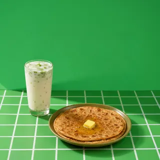 A plate of parathas topped with a melting cube of butter served alongside a glass of buttermilk on a green grid surface.
