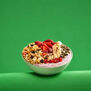 A smoothie bowl topped with granola, sliced strawberries, banana slices, seeds, and pomegranate seeds, presented in a light green bowl against a solid green background.