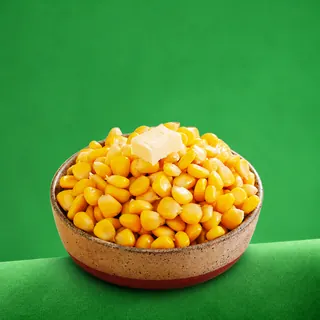 A bowl of steamed sweet corn kernels topped with a pat of butter against a solid green background.