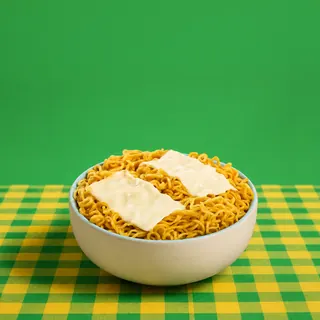 A bowl of instant noodles topped with two slices of cheese, placed on a green and yellow checkered tablecloth against a solid green background.