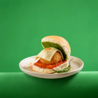 A cheese vada pav served on a white plate with a fried green chili against a solid green background.