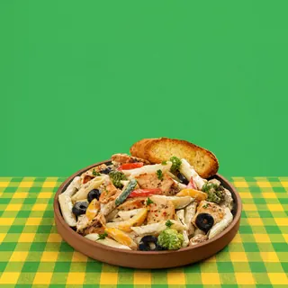 A plate of creamy chicken penne pasta with vegetables and garlic bread, served on a yellow and green checkered tablecloth against a solid green background.