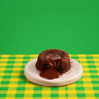 A chocolate lava cake with molten chocolate oozing out sits on a small round plate over a yellow and green checkered tablecloth against a green background.