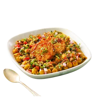A bowl of aloo tikki chaat topped with chickpeas, sev, chopped onions, and tomatoes, shown next to a spoon on a solid grey background.