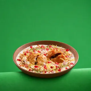 A bowl of dahi vada garnished with yogurt, sev, spices, and pomegranate seeds, set against a solid green background.