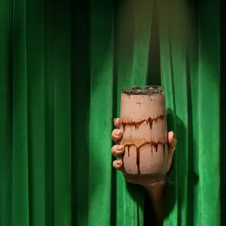 A hand holds a glass of chocolate milkshake decorated with chocolate syrup and cookie crumbs, emerging from behind green curtains.