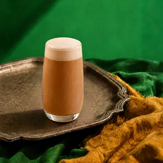 A tall glass of frothy cold coffee sits on a vintage metal tray against a green and gold fabric background.