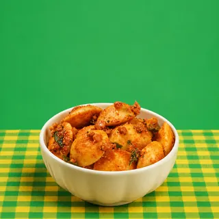 A white bowl filled with mini podi idlis coated in spices and curry leaves, set on a green and yellow checkered tablecloth against a solid green background.