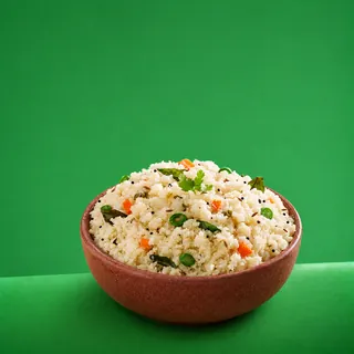 A brown bowl filled with upma garnished with curry leaves, carrots, and green chilies, set against a solid green background.